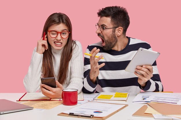 Indoor shot of angry man shouts at female trainee, holds touchpad, make project work together, have consultancy. Irritated young woman plugs ears, hears remarks from boss, isolated over pink wall