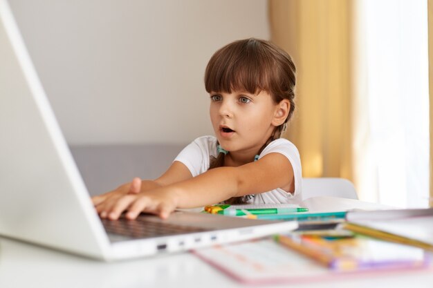 Снимок изумленного ребенка женского пола с косами, сидящего перед компьютером с очень удивленным выражением лица, с удивленным выражением лица смотрящего на дисплей ноутбука, онлайн-образование.