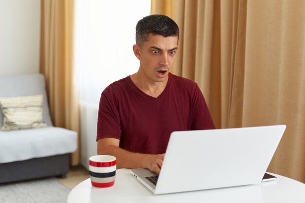 Indoor shot of astonished male freelancer working online, looking at notebook display with widely opened mouth, having system error, being scared to loose done work.