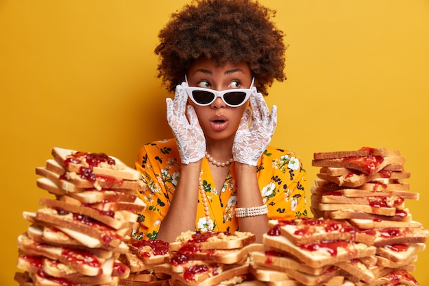 Indoor shot of astonished surprised dark skinned female model looks aside wears sunglasses and lace gloves sees something shocking isolated on yellow wall dressed in stylish clothes with jewelry