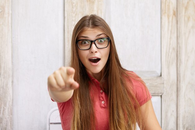 Indoor shot of astonished woman wearing polo shirt and rectangular glasses pointing her finger looking with surprised or shocked expression, mouth wide open.
