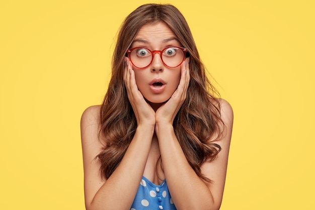 Indoor shot of astonished young woman with glasses posing against the yellow wall