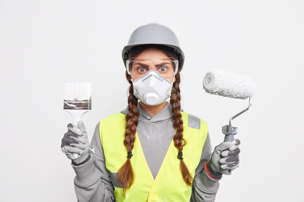 Indoor shot of attentive busy woman builder with pigtails looks stunned through transparent safety glasses holds paint brush and roller involvd in reconstruction wears protective helmet and face mask