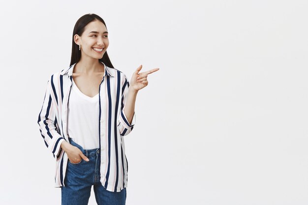 Indoor shot of attractive beautiful european female employee pointing and looking right with broad satisfied smile, holding hand in pocket, standing in confident pose, standing over gray wall