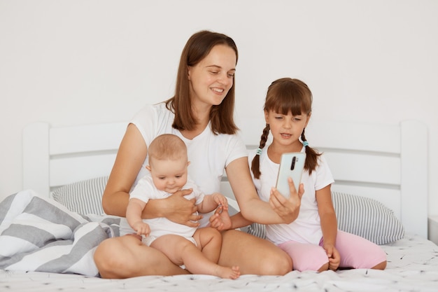 Indoor shot of attractive Caucasian female wearing white casual style t shirt sitting on bed with her two daughters, holding her infant baby in arms, posing with cell phone in hands.