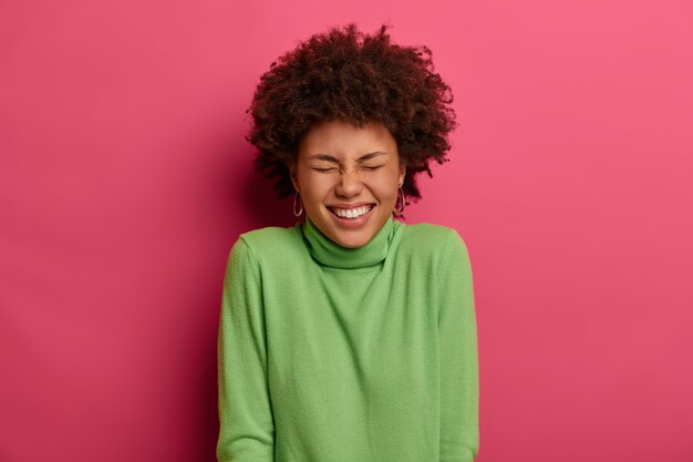 매력적인 곱슬 젊은 여성의 실내 촬영은 넓게 미소 짓고 하얀 치아를 보여주고 재미있는 농담을 웃으며 성실한 감정을 표현하고 분홍색 벽 위에 포즈를 취합니다. Peope, 감정, 라이프 스타일 컨셉