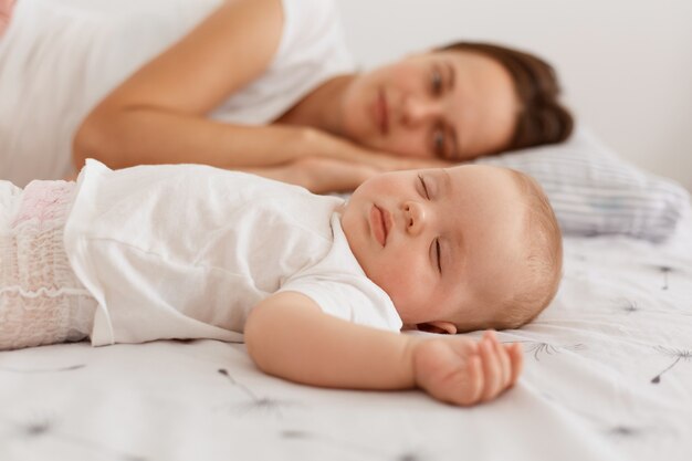 乳児と一緒にベッドに横になっている白いTシャツを着て、一緒に休んでいる魅力的な黒髪の女性の屋内ショット。ママはかわいい娘を見ています。
