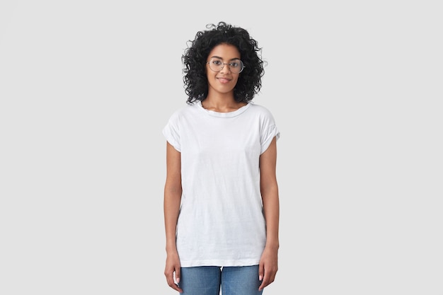 Indoor shot of attractive dark skinned female with crisp hair, wears round glasses, dressed in casual white t-shirt and jeans, poses indoor, has Afro hairstyle.