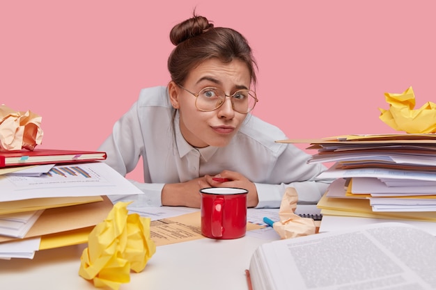 Indoor shot of attractive European student has hair knot, wears spectacles for good vision