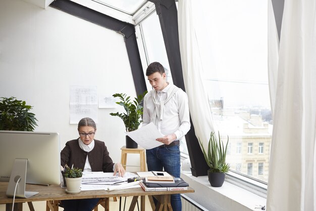 Indoor shot of attractive experienced mature woman architect sitting at desk and checking engineering drawings by her ambitious skilled young colleague. People, job, occupation and cooperation