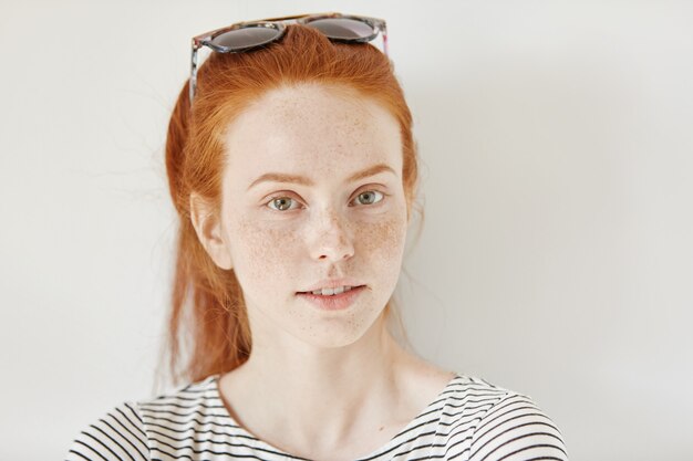 Indoor shot of attractive redhead woman model with freckles wearing trendy sunglasses on her head and sailor shot looking with faint smile