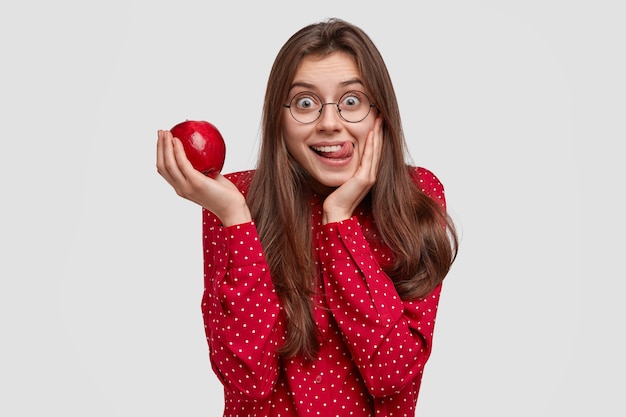 Indoor shot of attractive woman licks lips as holds fresh juicy apple, has fun, dressed elegantly, likes eating vitamins