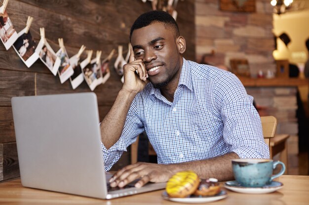 Indoor shot of attractive young dark-skinned employee in shirt using generic laptop computer