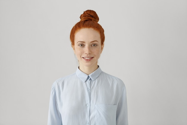 Indoor shot of attractive young redhead female with hair bun in shirt smiling happily