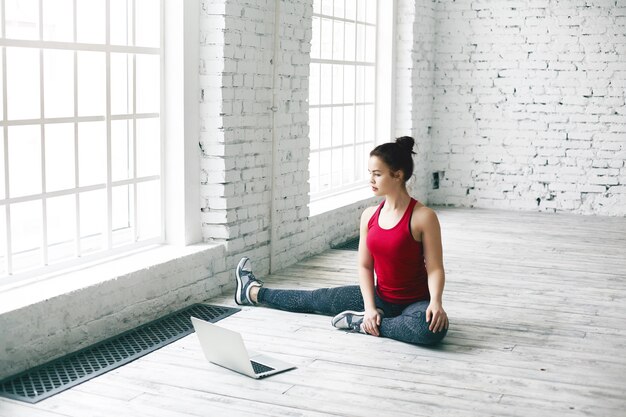Indoor shot of attractive young sportswoman doing yoga exercises and watching tutorial lesson on laptop computer at home, sitting on floor by large window wearing stylish sports clothes and sneakers