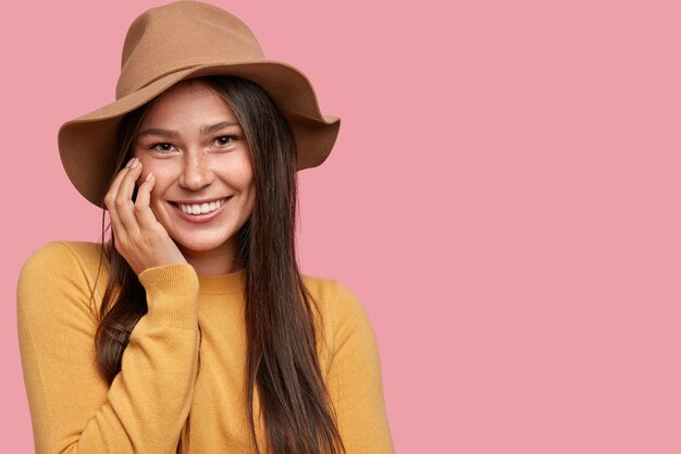 Indoor shot of attractive young woman with dark hair, smiles positively at camera, keeps hand on cheek, expresses happiness