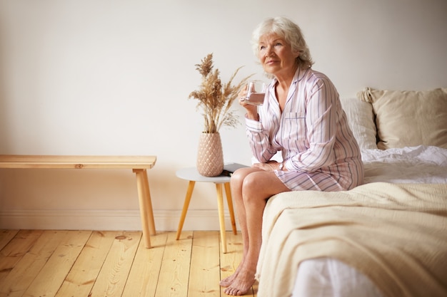 Free photo indoor shot of barefooted attractive gray haired female pensioner sitting on bed with feet on wooden floor, holding glass, drinking fresh water in morning. people, lifestyle, bedtime and aging concept