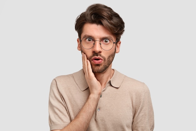 Indoor shot of bearded serious man with scared expression, touches cheek, has dark stubble, dressed in casual outfit, stands against white wall