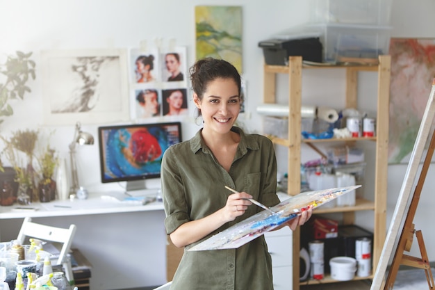 Indoor shot of beautiful brunette female painter wearing shirt, holding paint brush in hands standing near easel, creating masterpiece, smiling pleasantly while being glad to paint