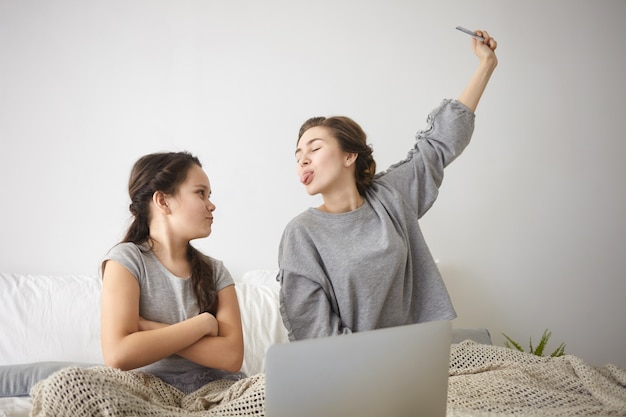 Indoor shot of beautiful casually dressed young female sitting on bed, teasing her angry annoyed daughter