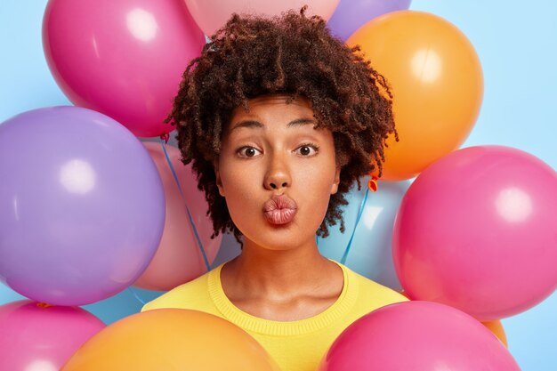 Indoor shot of beautiful curly haired dark skinned woman makes lips folded, has natural beauty, meets guests during her birthday, stands against inflated colorful balloons, dressed in yellow jumper