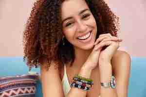 Free photo indoor shot of beautiful dark skinned mixed race young female with afro hairstyle has pleasant smile, poses on sofa, wears bracelet, hears funny story from friend