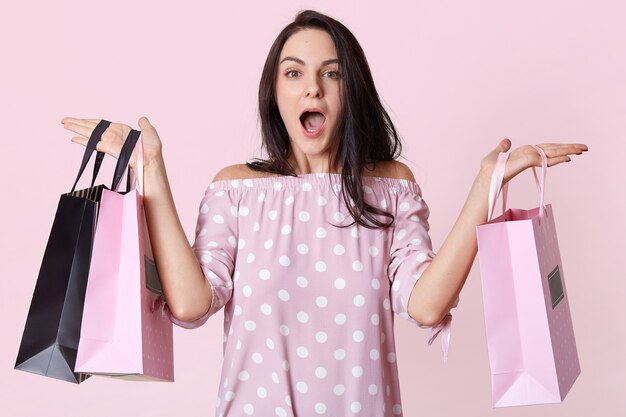 Indoor shot of beautiful fashionable Caucasian woman holding shopping bags in both hands, has shocked facial expression