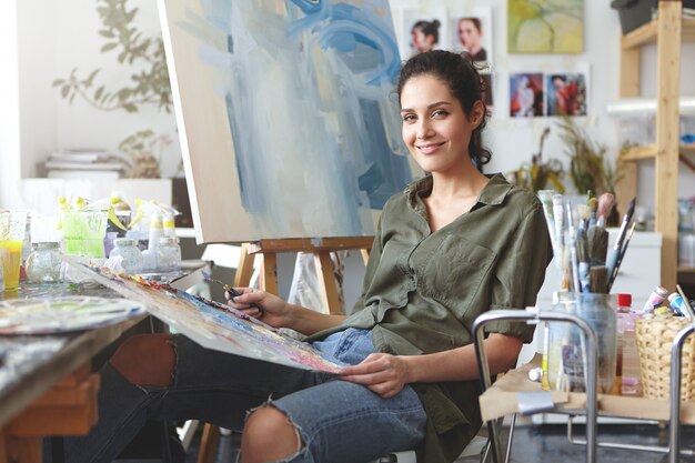 Indoor shot of beautiful female painter wearing shirt and jeans, sitting at chair, mixing colorful oils, making brush of strokes on easel. Female art-lover practicing drawing at her workshop