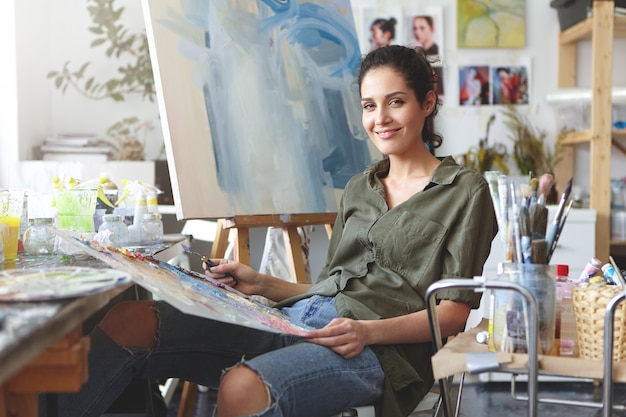 Indoor shot of beautiful female painter wearing shirt and jeans, sitting at chair, mixing colorful oils, making brush of strokes on easel. Female art-lover practicing drawing at her workshop