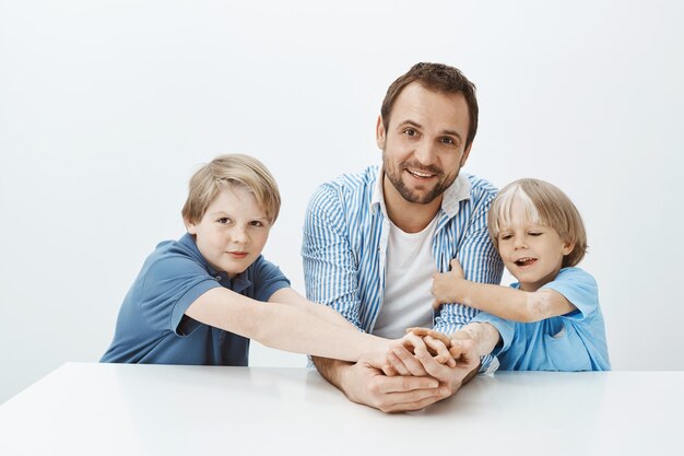 Indoor shot of beautiful happy dad and sons sitting at table and smiling broadly, holding hands and gazing with broad smile, fooling around