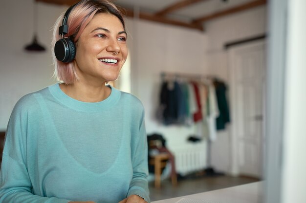 Indoor shot of beautiful happy student girl in blue sweatshirt using wireless headphones, having online examination, sitting at home. People, education, learning, technology and electronic gadgets