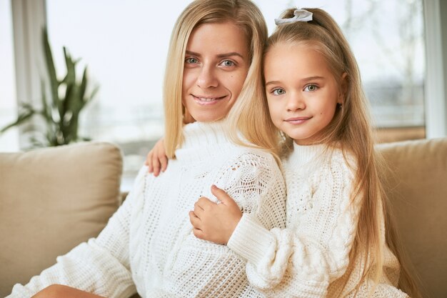 Indoor shot of beautiful little girl embracing her attractive young female keeping hands around her waist, both dressed in cozy warm sweaters,  with joyful happy smiles, having fun