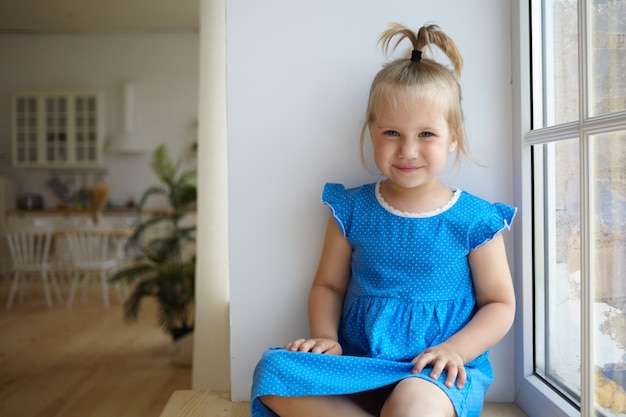 Tiro al coperto di bella bambina in età prescolare seduta sul davanzale della finestra con coda di cavallo divertente, vestita in abito blu, sorridendo felicemente alla telecamera, avendo un aspetto spensierato, cucina moderna in background