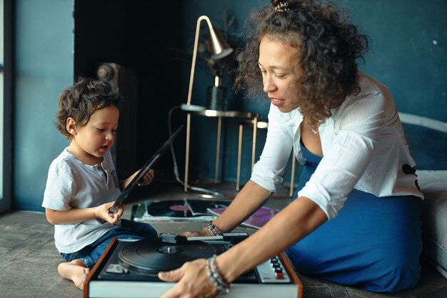 Indoor shot of beautiful mixed race melomaniac female sitting on floor with her little son using turntable, listening to music together