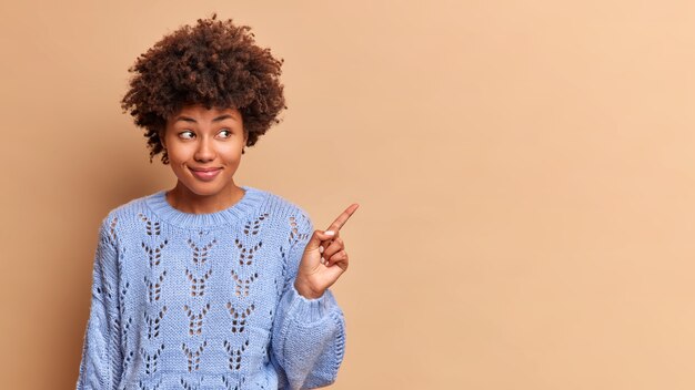 Indoor shot of beautiful smiling woman points right on blank space promots product or advertises company banner gives recommendation dressed in casual sweater isolated over beige wall