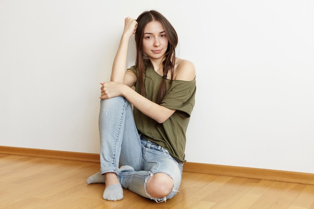 Free photo indoor shot of beautiful teenage woman with messy hair wearing ragged jeans and stylish oversize top sitting on floor in her white bedroom, spending weekend at home and waiting for friends to come