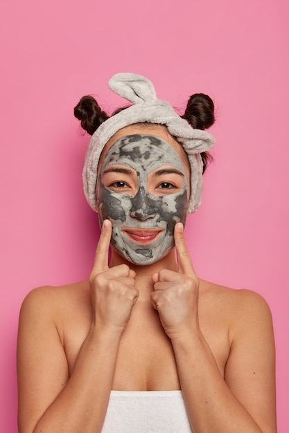 Indoor shot of beautiful woman applies nutrient cleansing peel off face mask, indicates with both fore fingers at cheeks, cares about appearance, gets beauty procedures in spa salon, wrapped in towel