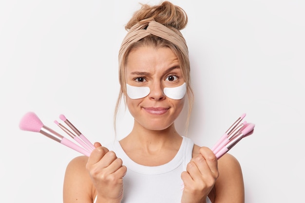 Indoor shot of beautiful woman enjoys beauty day holds cosmetic brushes for applying makeup applies moisturizing patches under eyes isolated over white background refreshes her skin. Daily routine