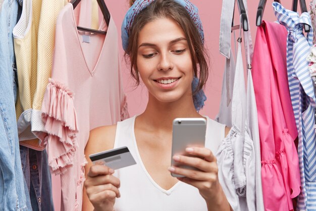Indoor shot of beautiful woman with gentle smile, standing near variety of clothes being shopaholic, buying garment online, using smart phone and credit card. People, shopping, clothes concept