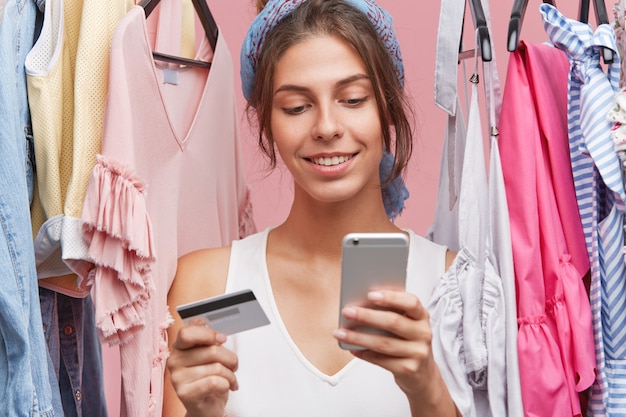 스마트 폰 및 신용 카드를 사용 하여 온라인 쇼핑 의류 구매 shopaholic 되 고 다양 한 옷 근처에 서 부드러운 미소와 아름 다운 여자의 실내 샷. 사람, 쇼핑, 옷 개념
