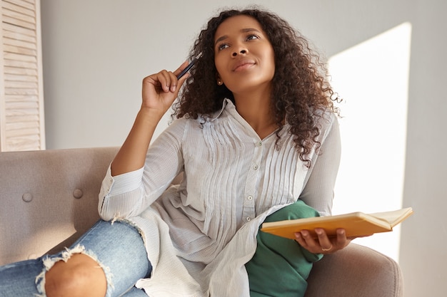Indoor shot of beautiful young Afro American lady in blouse and ripped jeans having pensive facial expression, looking up, scratching head with pen, making notes in copybook, developing business plan