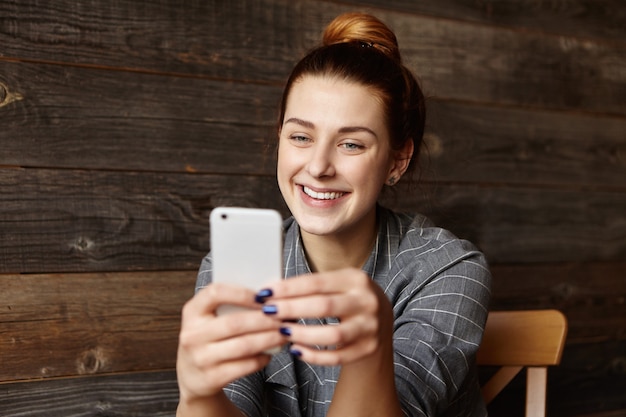 Крытый снимок красивой молодой женщины, весело улыбаясь, переписывая текстовые сообщения друзьям