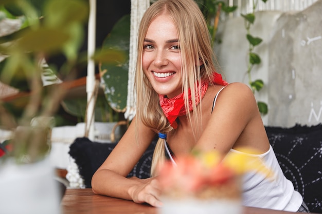 Indoor shot of blonde female with charming shining smile, wears bandana on neck, sits on comfortable sofa, rests at coffee shop