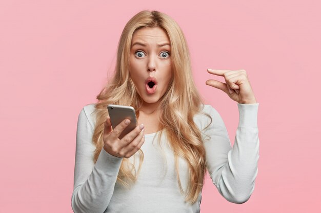 Снимок красивой блондинки с голубыми глазами, небрежно одетой, держит смартфон, проверяет онлайн-счет, изолированную над розовой стеной. Шокированная женщина с привлекательной внешностью моделей в