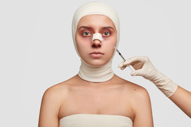 Indoor shot of bruised woman with pale skin, has plaster on nose, wrapped in bandages, recieves injection from surgeon, has serious facial expression.