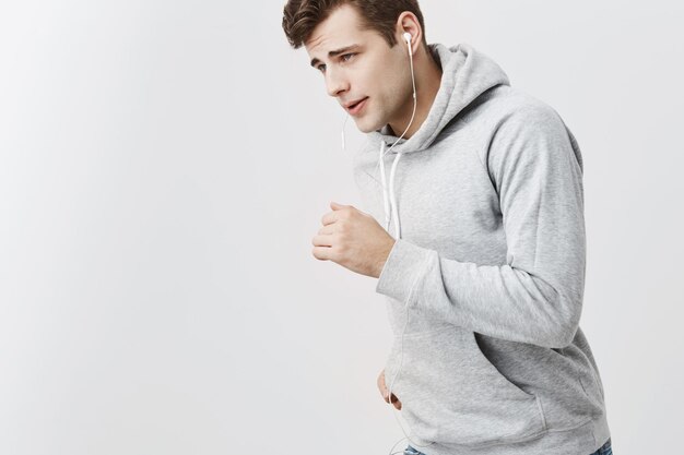 Indoor shot of brunnete handsome man with blue eyes in gray hoodie and jeans with earphones running against gray studio wall