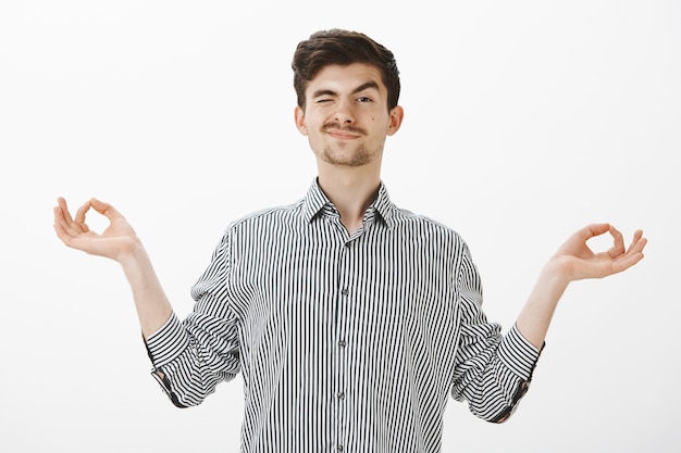 Indoor shot of carefree good-looking friendly guy with moustache in shirt, spread hands in zen gesture, peeking with one eye and smiling while watching students during practicing yoga or meditation