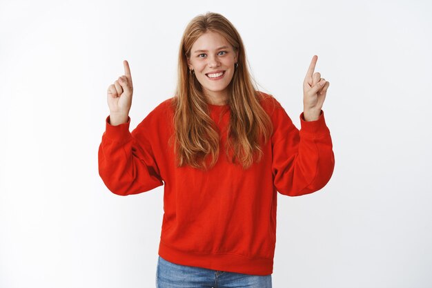 Indoor shot of carefree happy young charismatic fair-haired woman with freckles and blue eyes smiling sincere and happily standing in cute red oversized sweater pointing up with delighted expression