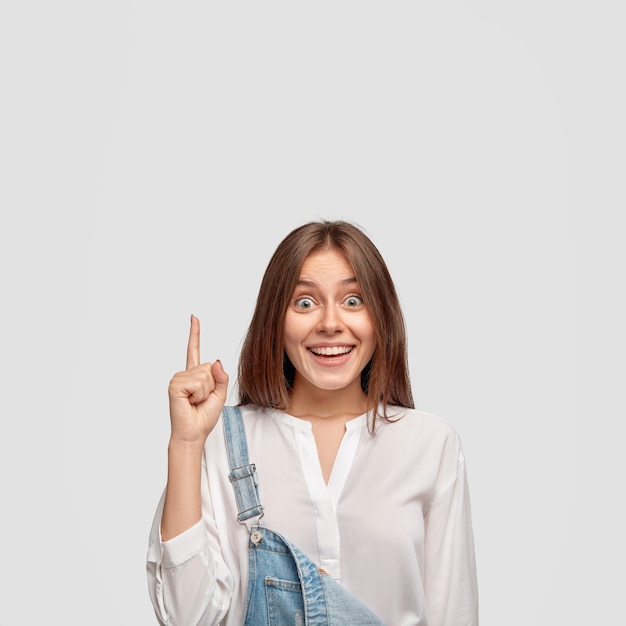 Free photo indoor shot of carefree satisfied brunette woman with pleasant cheerful expression points upwards with index finger