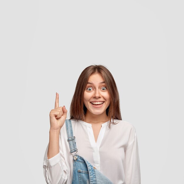 Indoor shot of carefree satisfied brunette woman with pleasant cheerful expression points upwards with index finger